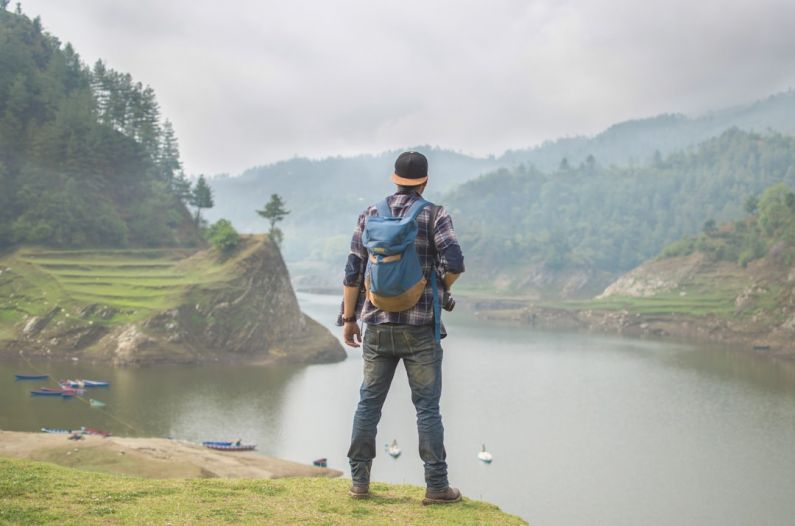 Traveler - person in blue denim pants carrying blue backpack in front of body of water during daytime