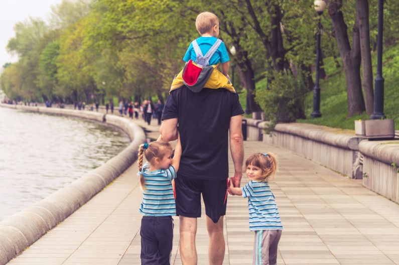 Family Fun - man in black t-shirt and brown shorts holding girl in blue and black jacket walking