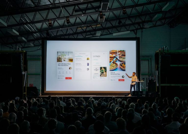 Event - person discussing while standing in front of a large screen in front of people inside dim-lighted room